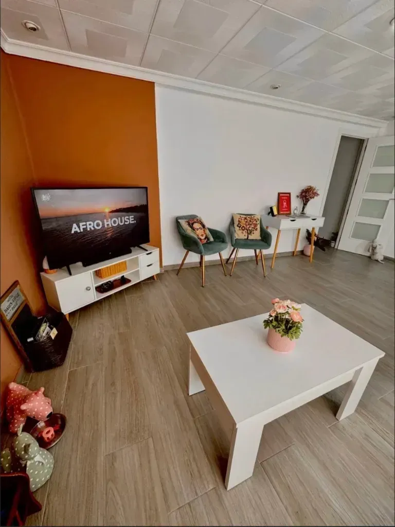 Sala de estar moderna con pared decorativa de color naranja, televisor, mesa de centro blanca y dos sillas junto a una estantería.