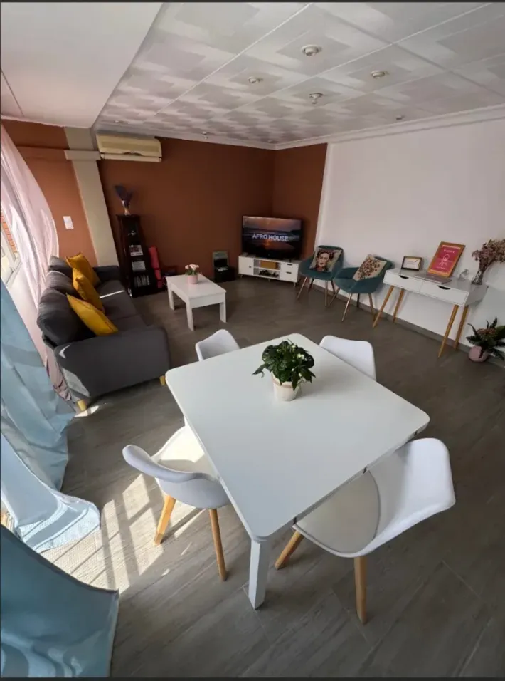Sala de estar luminosa con mesa de comedor blanca, sillas, sofá, televisor y pared decorativa marrón.