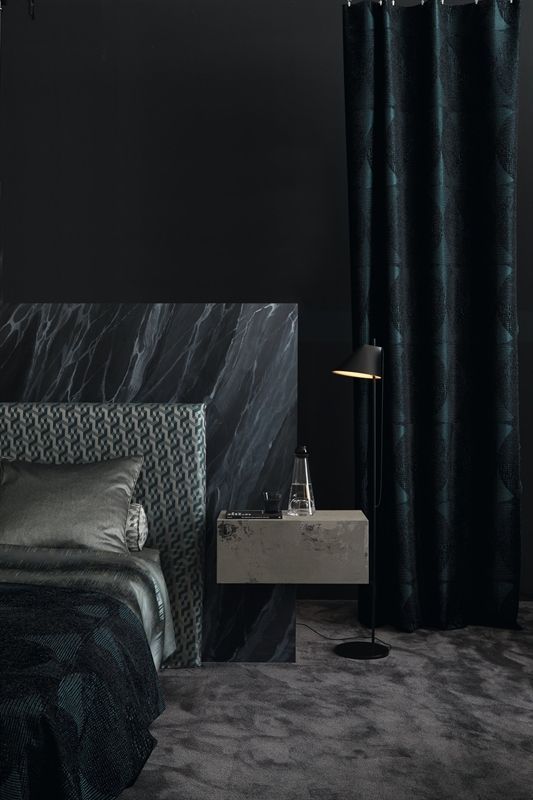 Ein Schlafzimmer mit Bett, Nachttisch, Lampe und schwarzen Vorhängen.