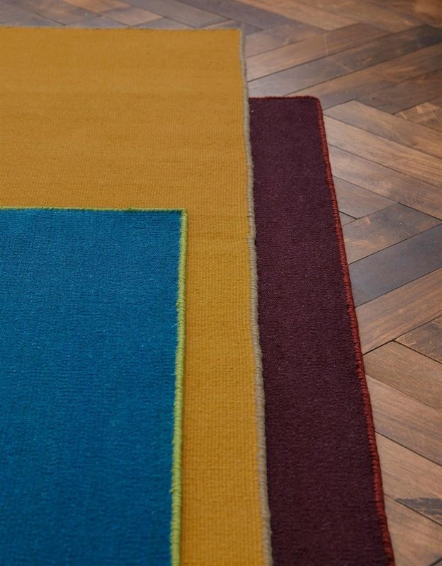 Auf einem Holzboden sind drei verschiedenfarbige Teppiche übereinander gestapelt.