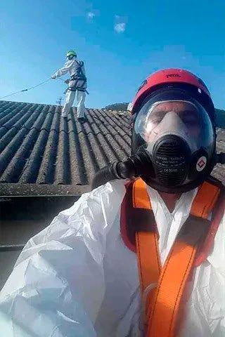 Una persona con casco rojo y equipo de protección se toma una selfie en un tejado, con un compañero de trabajo sujeto con una correa detrás.