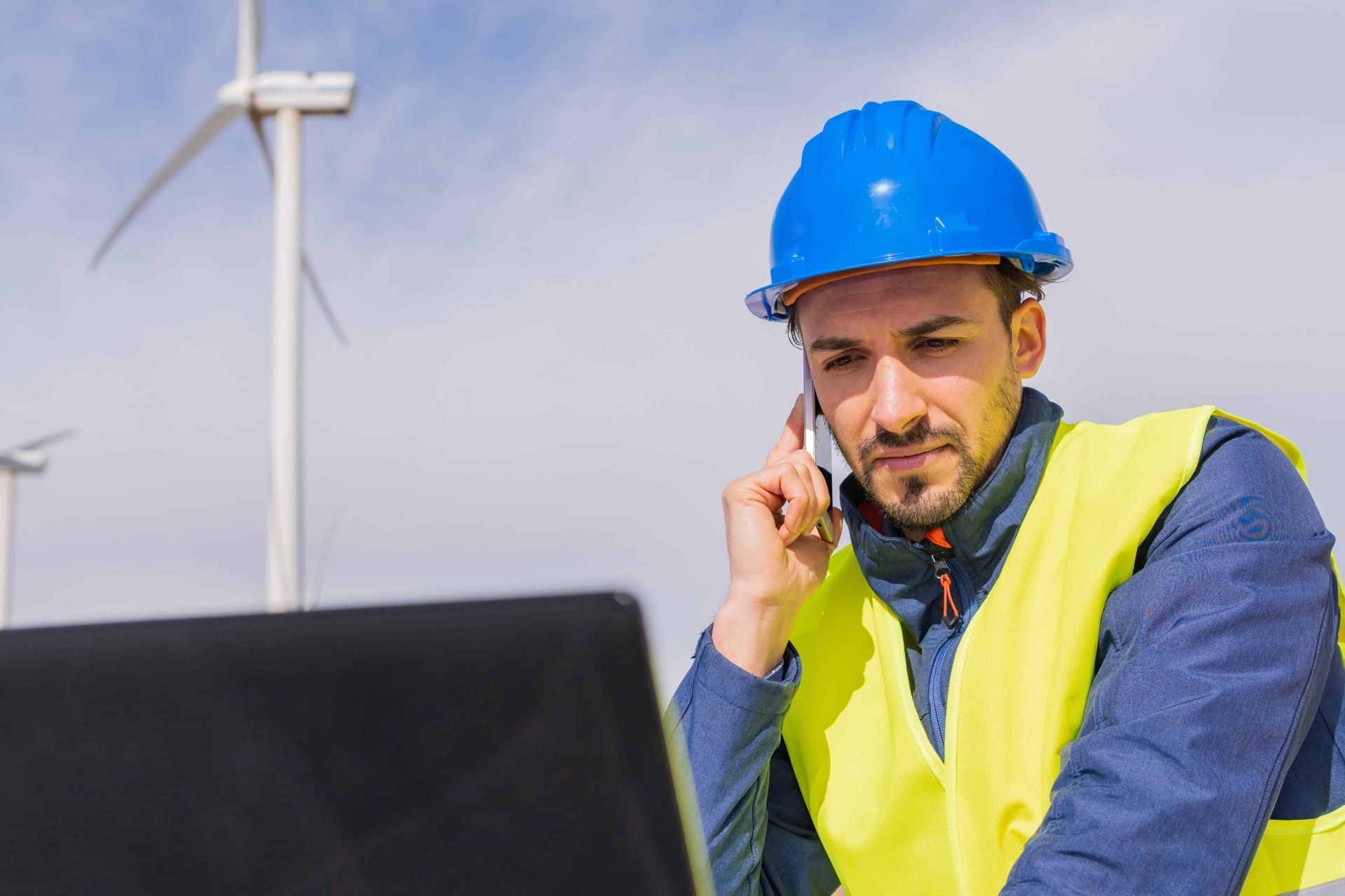 Un técnico con casco azul y chaleco reflectante habla por teléfono mientras trabaja en un ordenador portátil en un parque eólico.