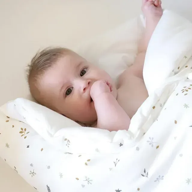 Un bebé está acostado sobre una manta blanca con la mano en la boca.