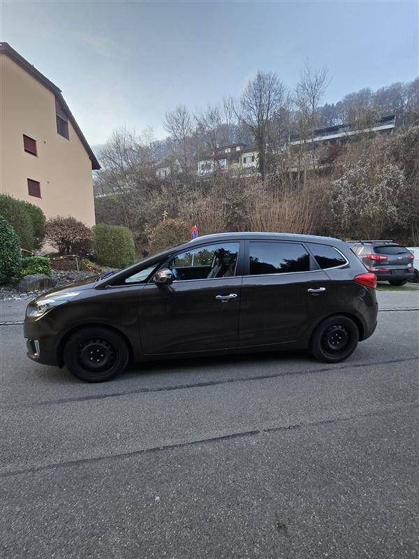 Ein dunkelbraunes Auto parkt auf einer Strasse; im Hintergrund sind ein Gebäude und ein Hügel zu sehen.