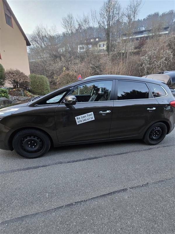 Ein brauner Kia Carens parkt auf der Strasse. Schwarze Felgen und ein Aufkleber an der Vordertür. Im Hintergrund Bäume.