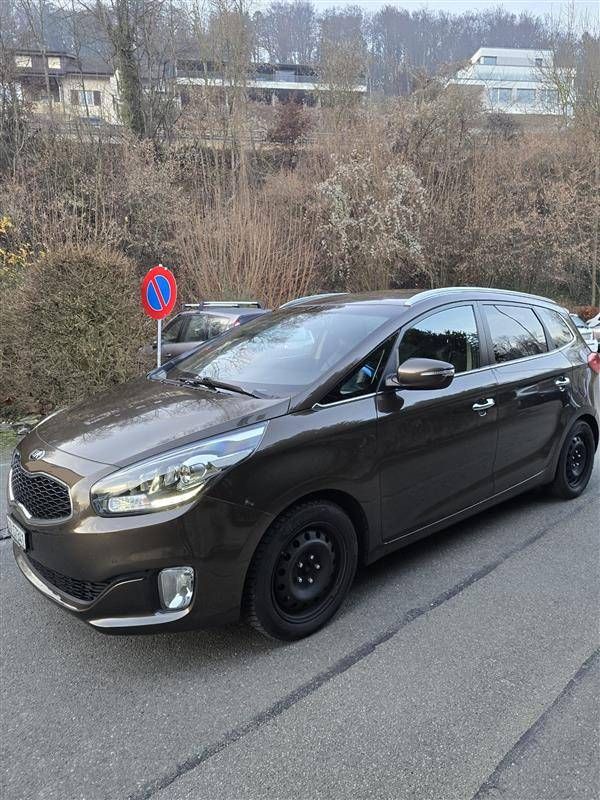 Ein brauner Kia Carens parkte auf einer Strasse in der Nähe von Bäumen und Gebäuden.