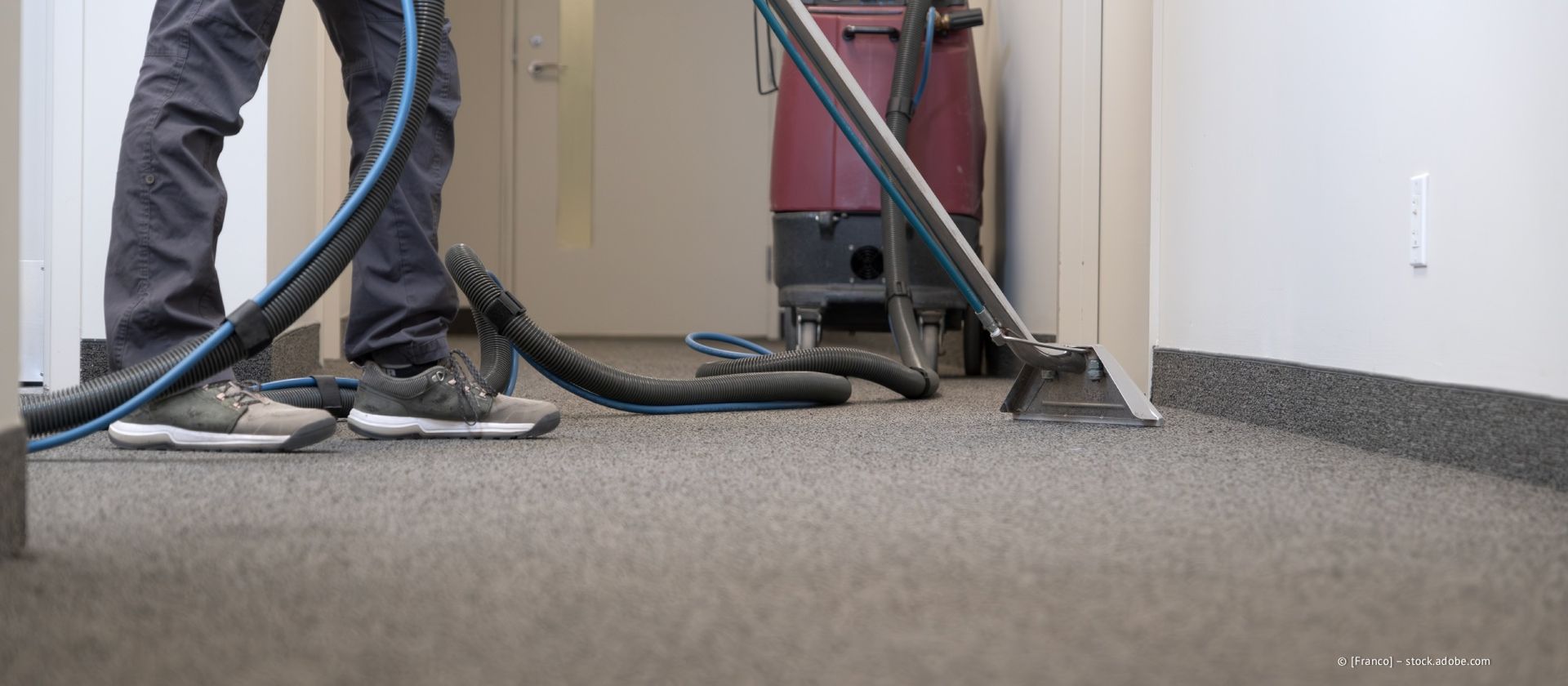 Person saugt mit einer professionellen Reinigungsmaschine einen grauen Teppich in einem Flur.