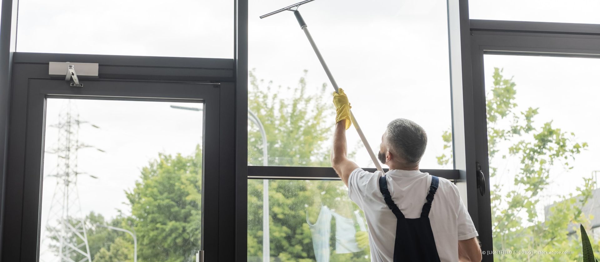 Ein Mann reinigt ein großes Fenster von der Innenseite eines Gebäudes aus. Er verwendet einen Abzieher mit langem Griff.