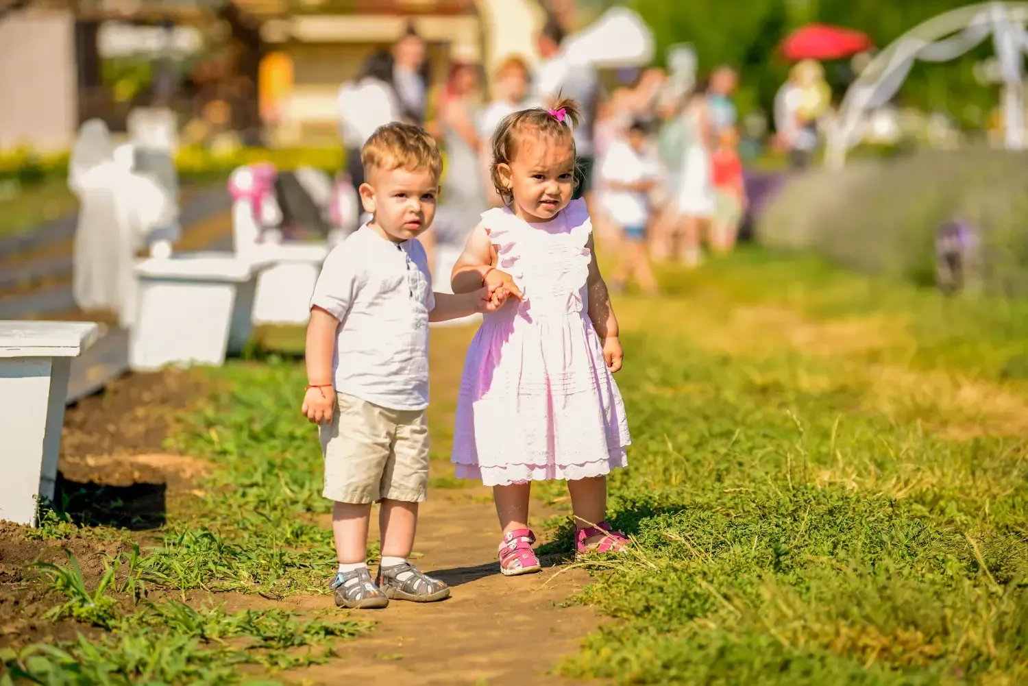Dos niños pequeños de pie en un sendero soleado del jardín, tomados de la mano entre flores y vegetación.