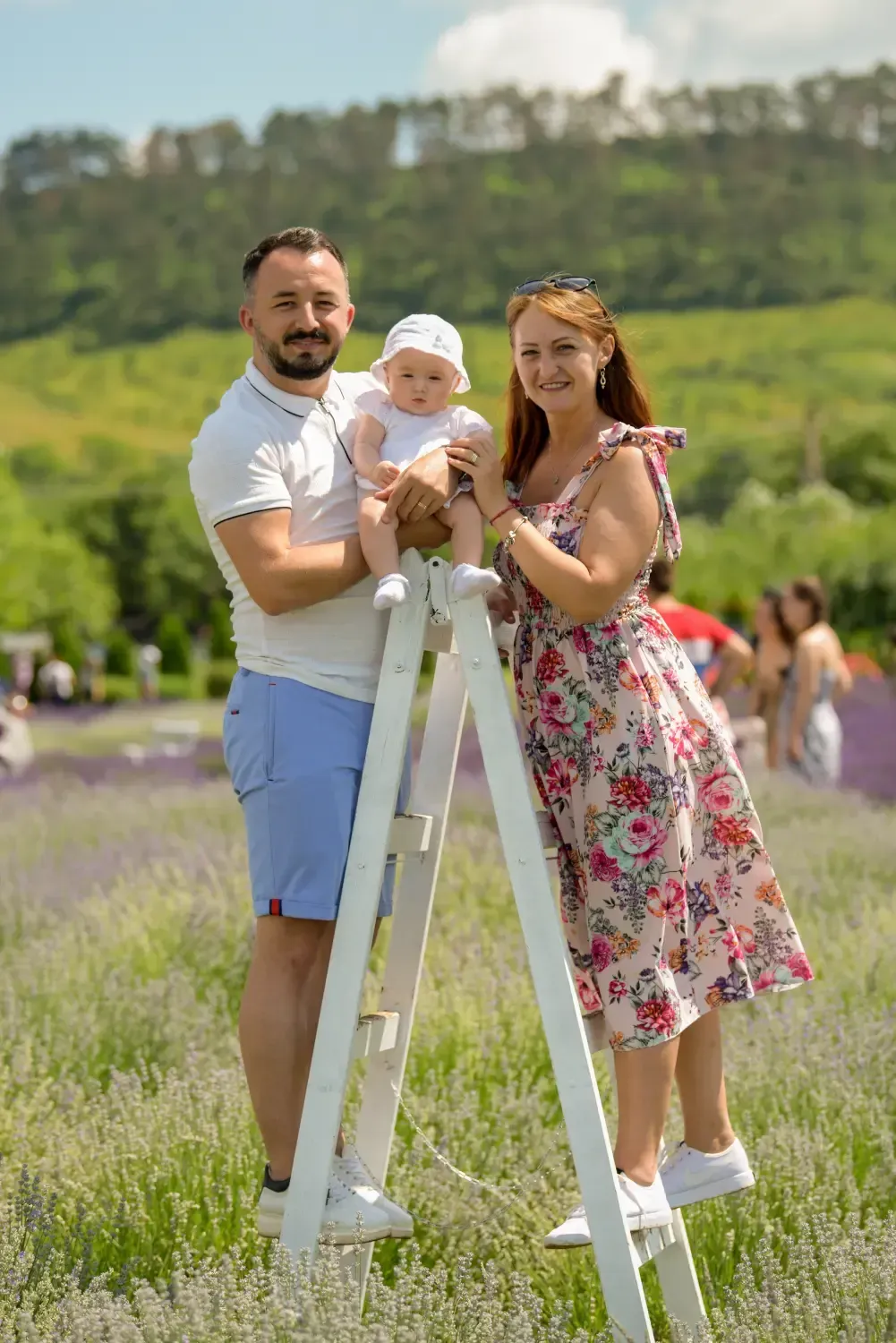 Familia posando sobre una escalera blanca en un campo soleado, con árboles y una multitud borrosa al fondo.