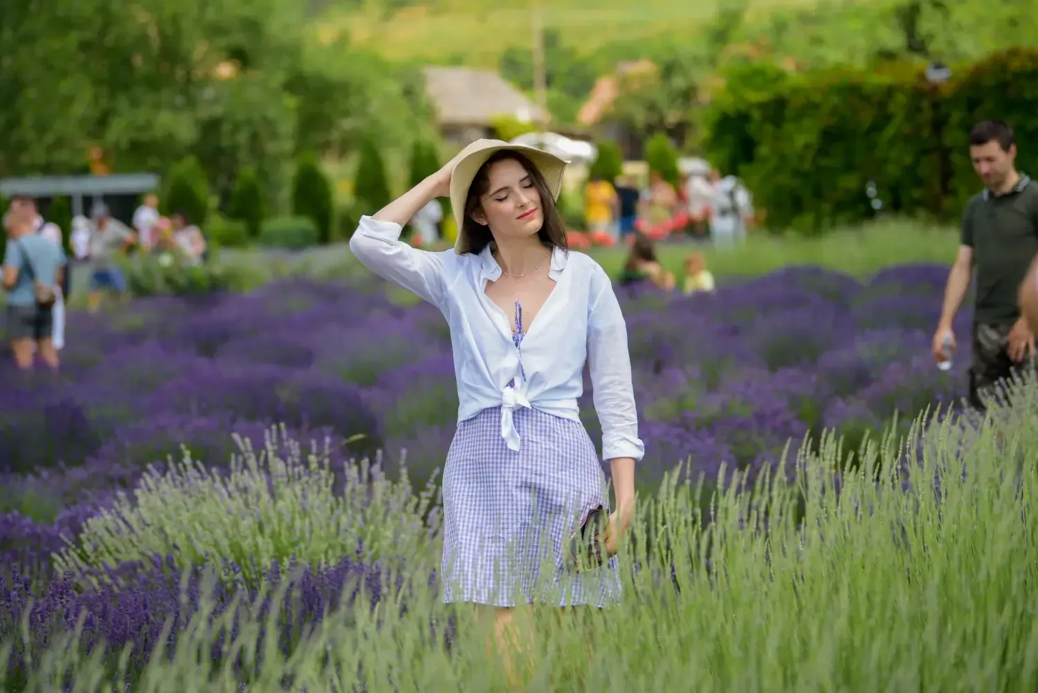 Mujer en un campo de lavanda, vestida con una camisa blanca y una falda a cuadros, sonriendo y sosteniendo un sombrero.