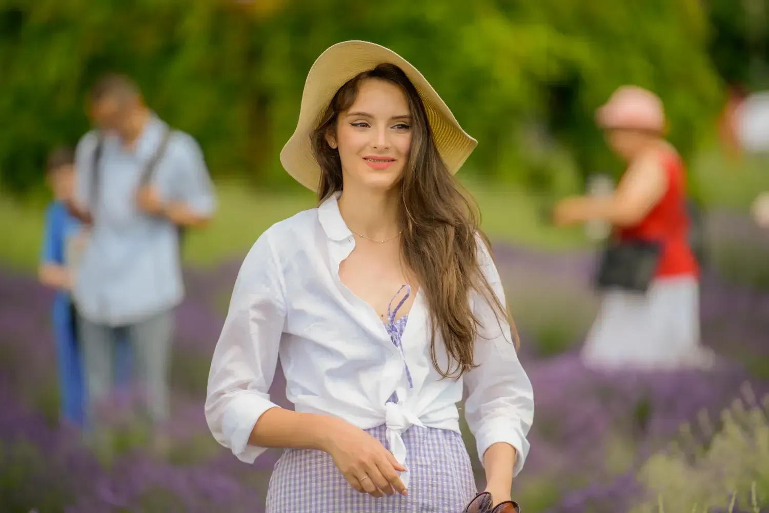 Mujer con camisa blanca y sombrero de pie en un campo de lavanda, con dos personas borrosas al fondo.