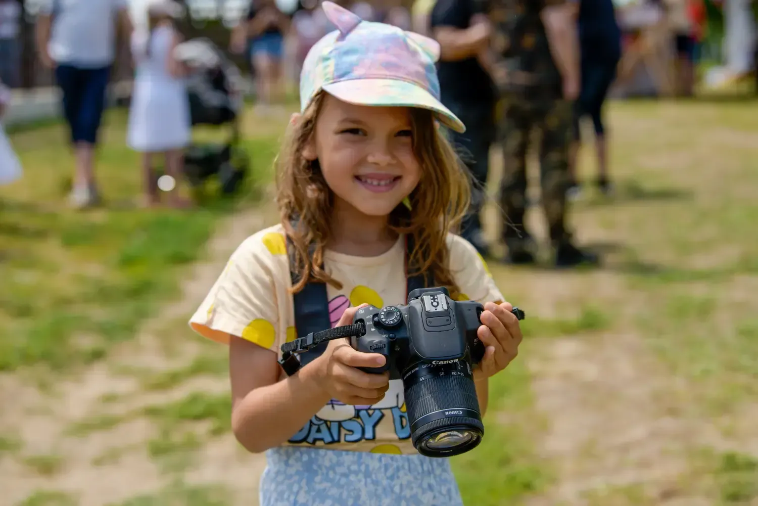 Un niño sonriente sostiene una cámara al aire libre en un parque con césped, con personas borrosas al fondo.