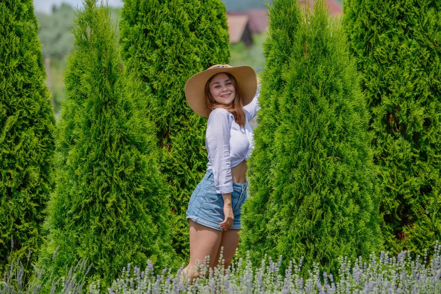 Una mujer con sombrero de paja, de pie entre altos arbustos verdes en un jardín, sonríe a la cámara.