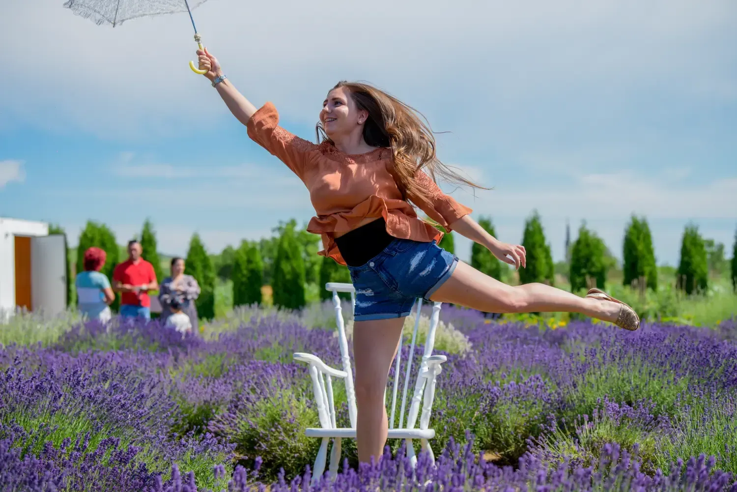 Mujer con camisa naranja haciendo equilibrio en un campo de lavanda, extendiendo la mano hacia una rama florida.