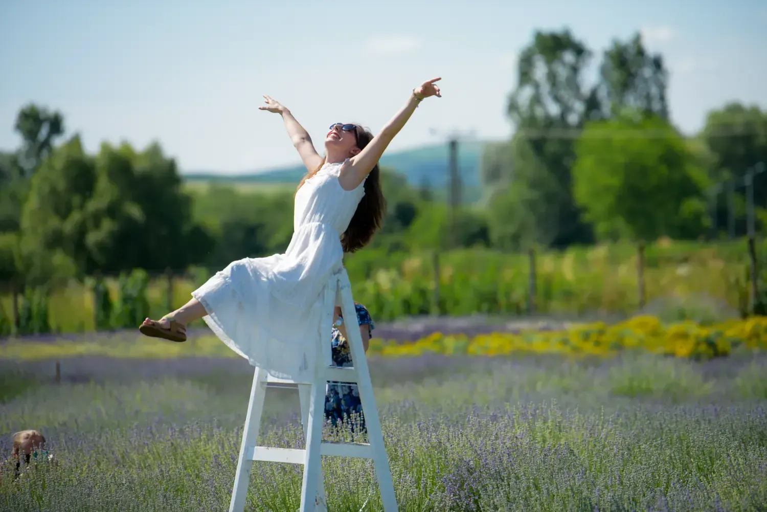 Mujer con vestido blanco estirándose en una escalera en un campo de lavanda bajo un cielo soleado.