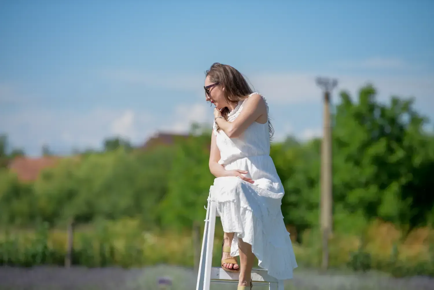 Mujer con vestido blanco sentada al aire libre en una silla en un campo soleado, mirando hacia abajo.
