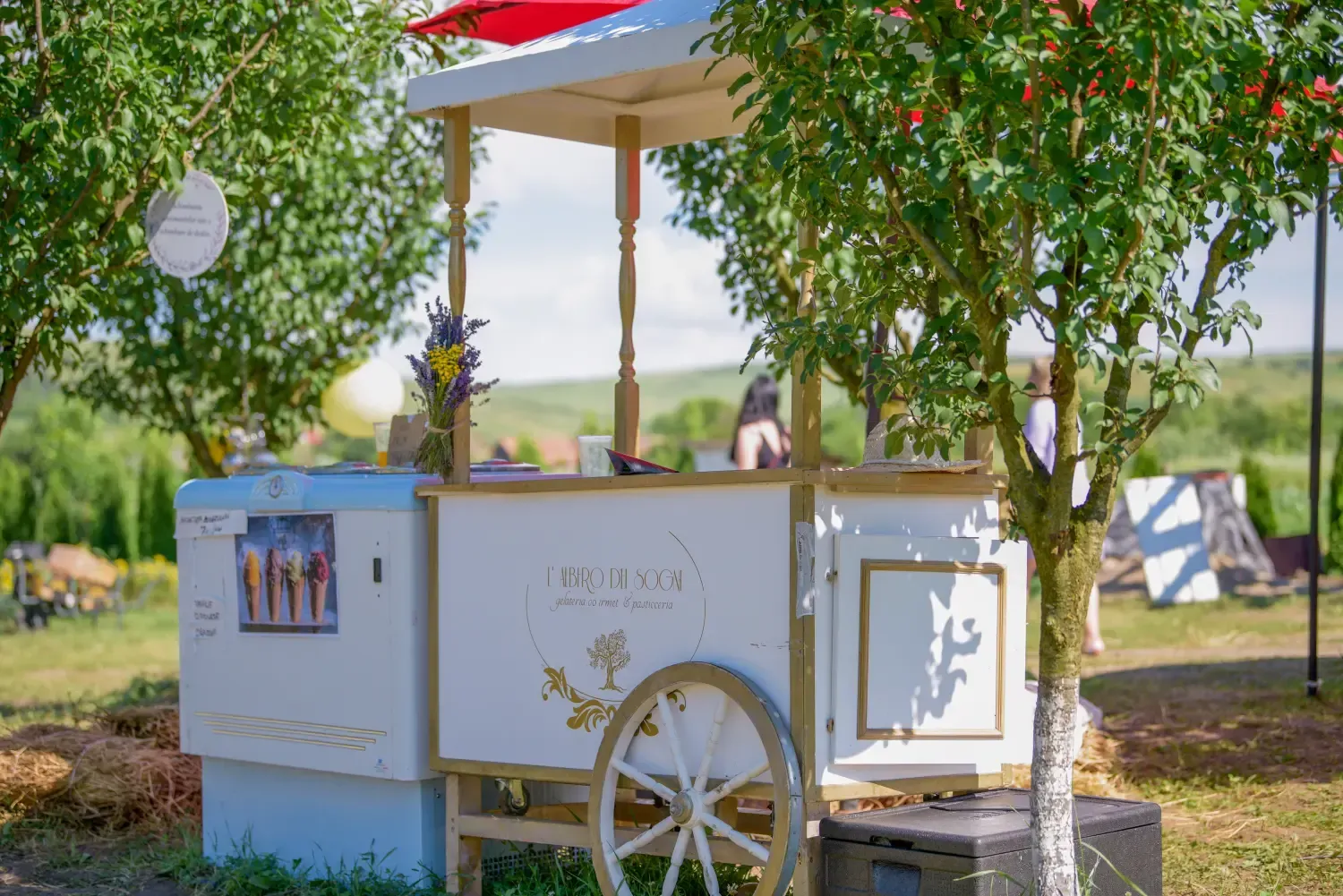 Un carrito de postres blanco bajo un toldo en un jardín soleado, enmarcado por plantas colgantes y una sombrilla roja.
