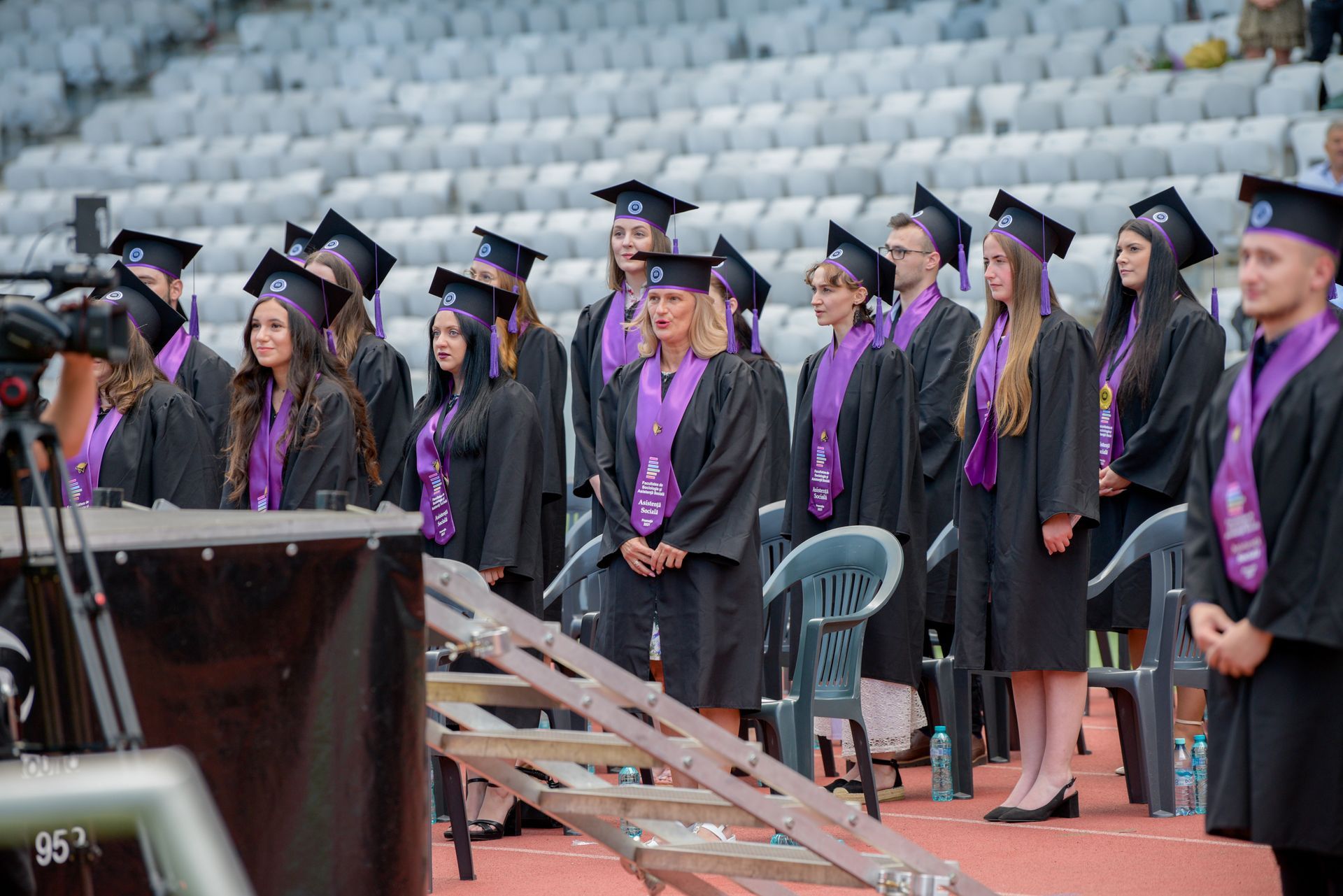 Los graduados, vestidos con birretes y togas negras y estolas moradas, permanecen de pie en la pista del estadio durante la ceremonia.