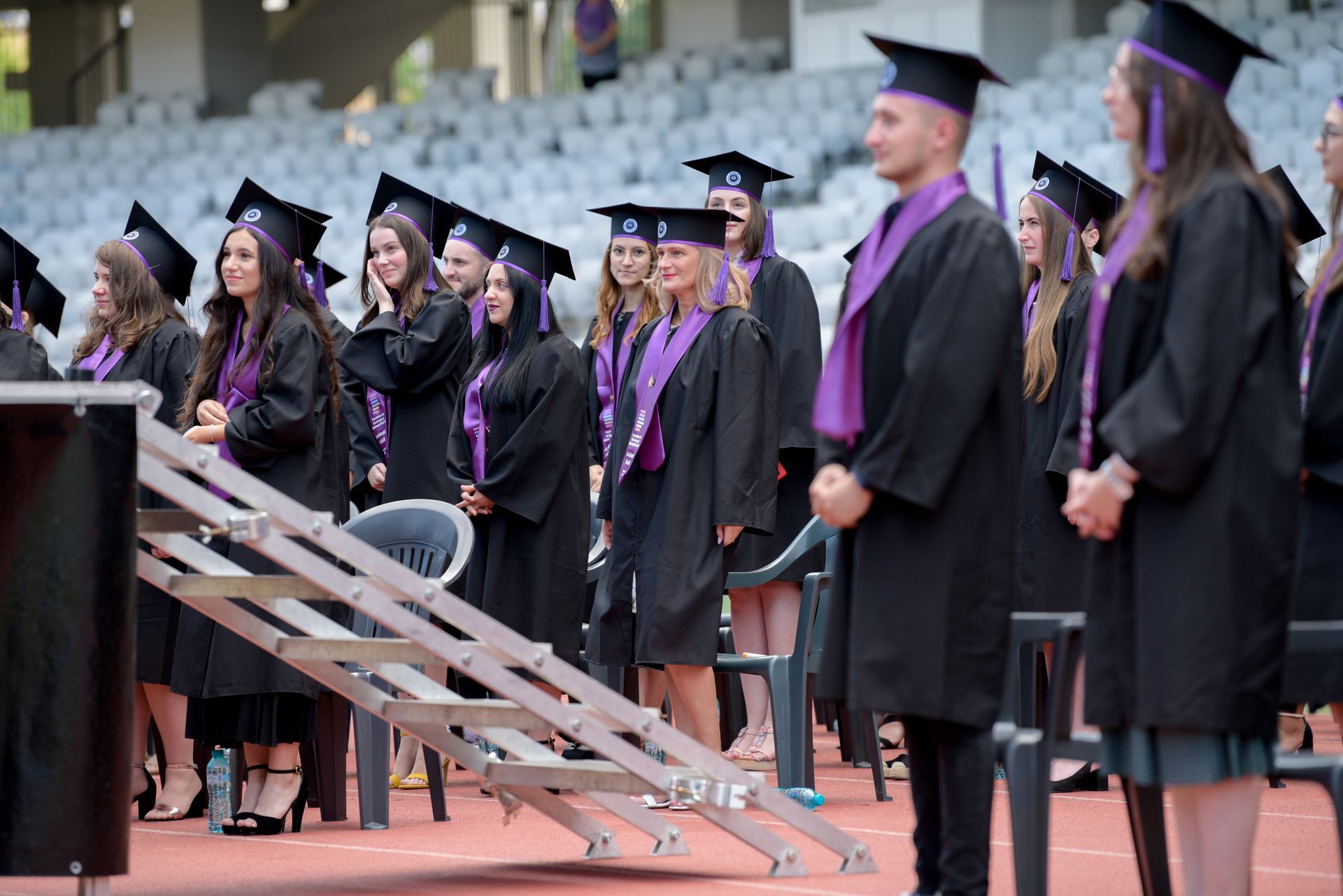 Los graduados, vestidos con togas negras y estolas moradas, permanecen de pie en la pista del estadio durante la ceremonia de graduación.