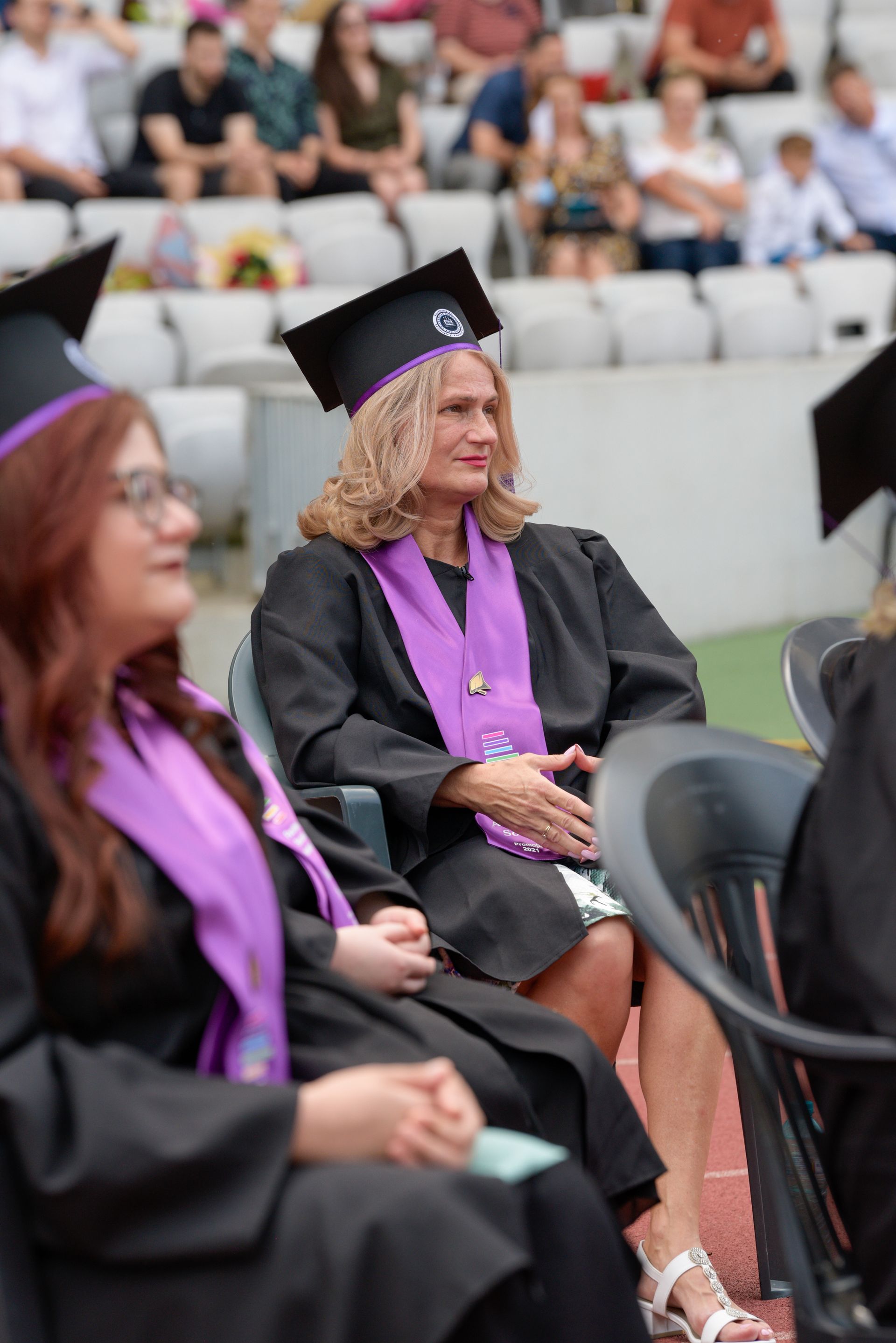Los graduados, vestidos con togas y birretes negros, se sientan en las gradas del estadio durante una ceremonia al aire libre.