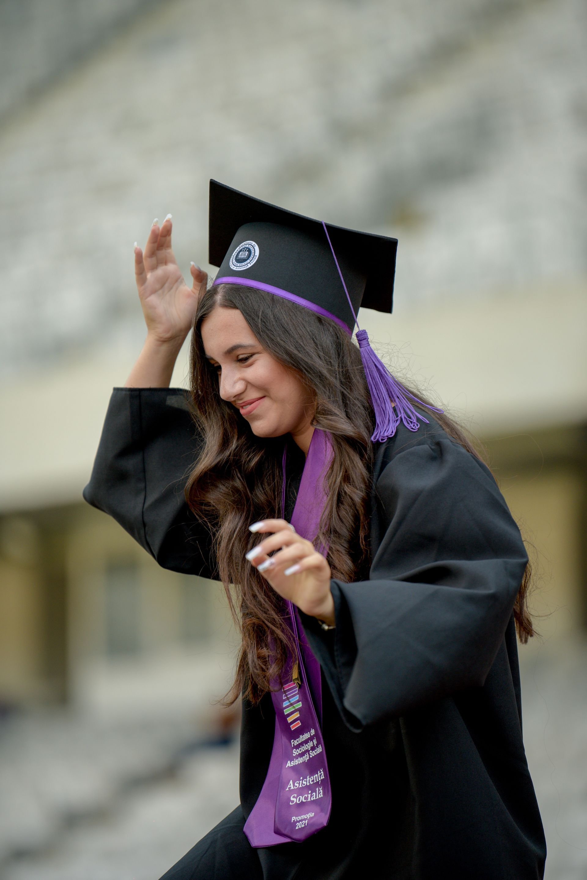 Graduado con toga y birrete negros ajustándose el birrete, sonriendo al aire libre con estola y borla moradas.