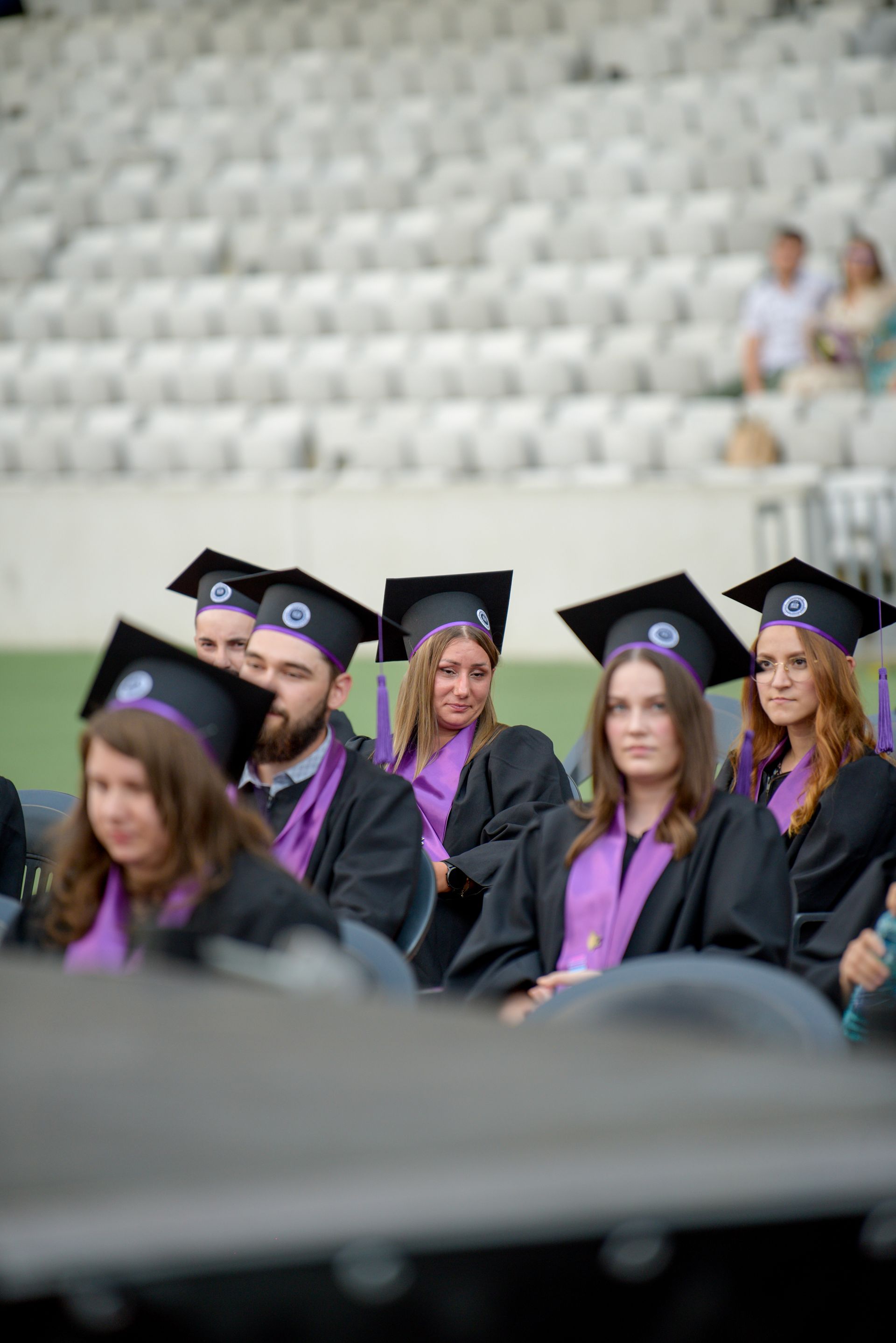 Los graduados, ataviados con togas y birretes negros, se sentaron en una ceremonia al aire libre; entre ellos, se podía ver a una mujer con una estola morada.