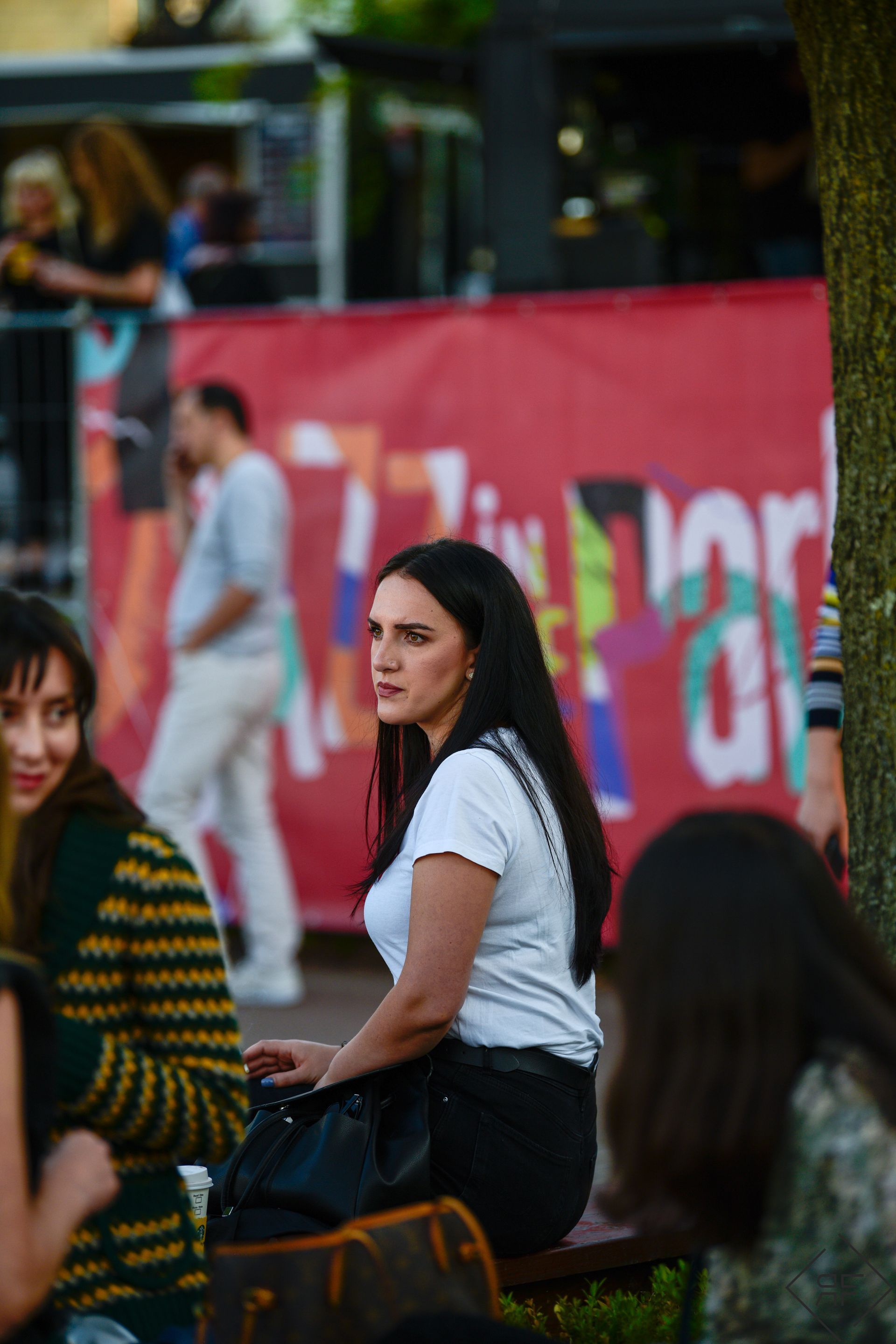 Mujer con camisa blanca sentada al aire libre cerca de una pancarta roja, mirando por encima del hombro.