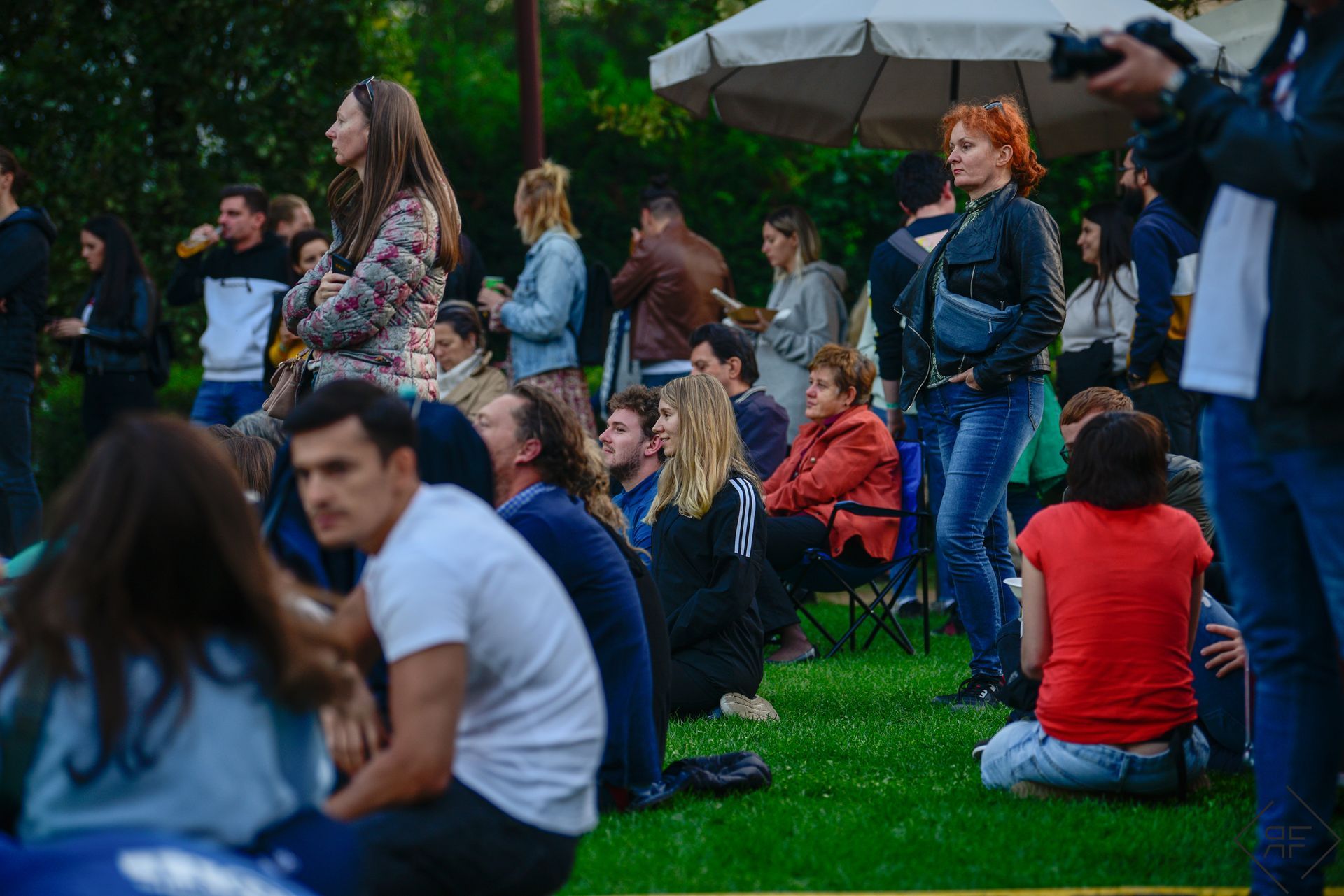 Multitud sentada sobre el césped en un evento al aire libre, con gente de pie fotografiando la escena.
