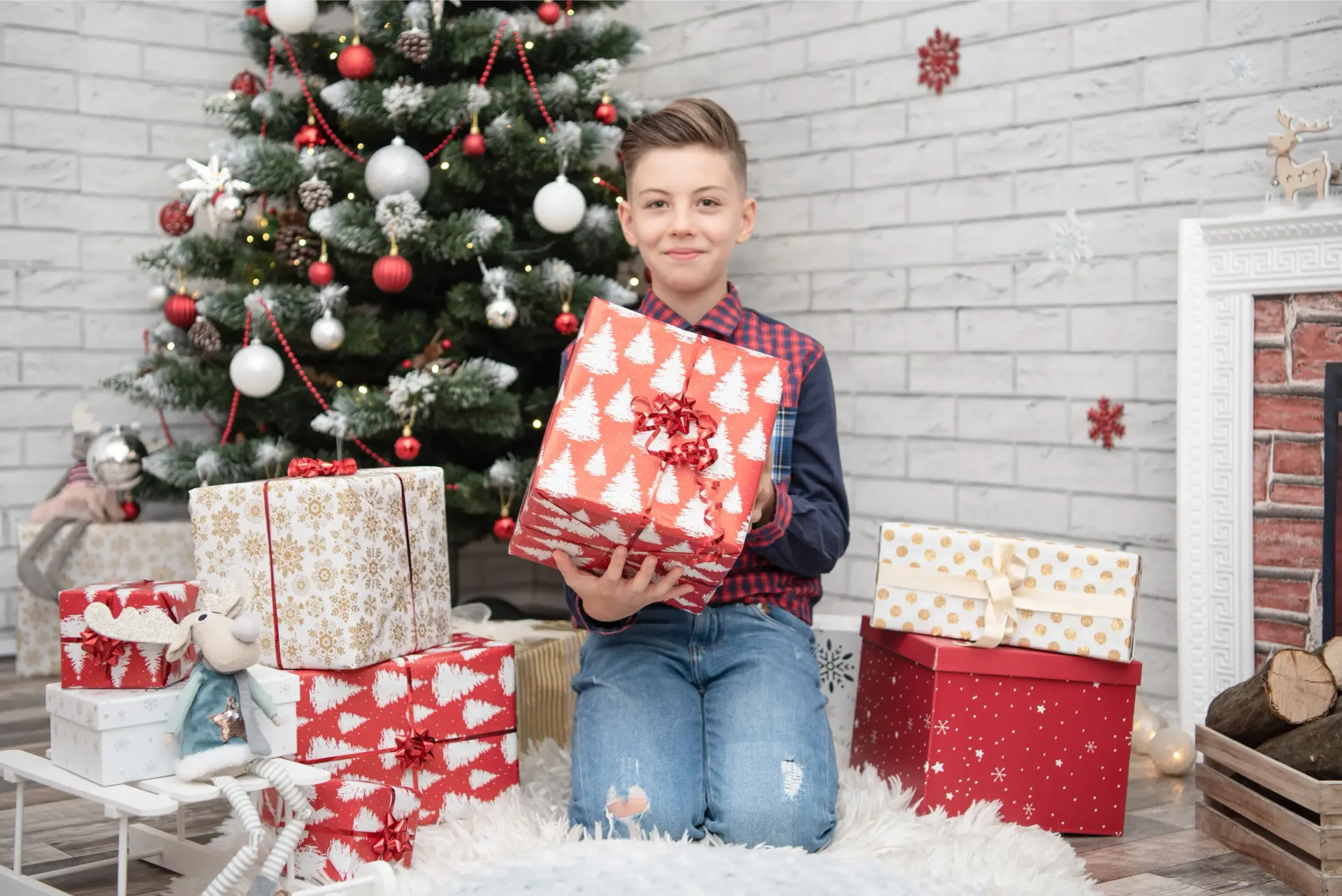 Niño arrodillado junto a un árbol de Navidad decorado, sosteniendo un regalo rojo en una escena navideña nevada.
