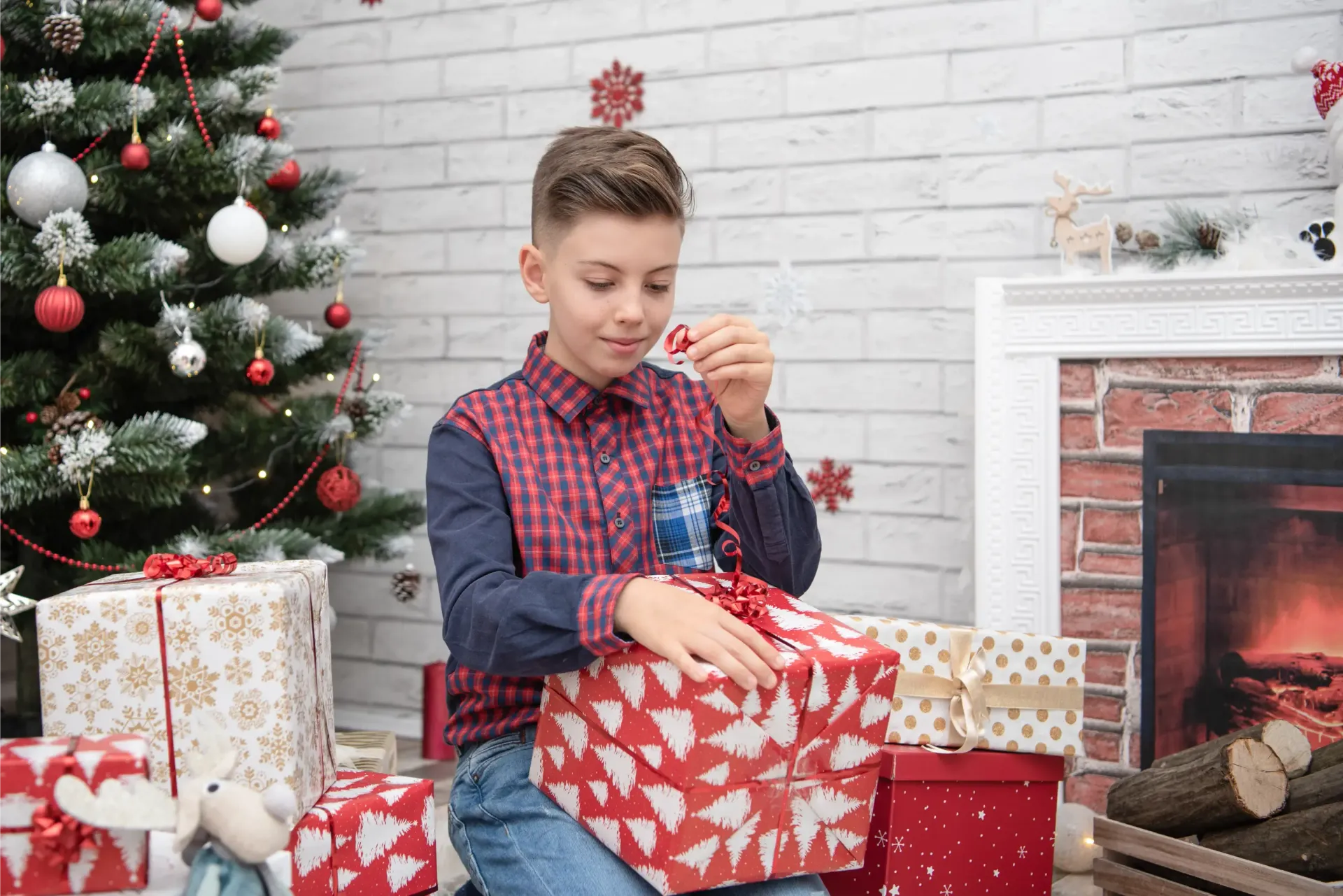 Un niño abre una caja de regalo roja y blanca junto a un árbol de Navidad decorado y una chimenea.