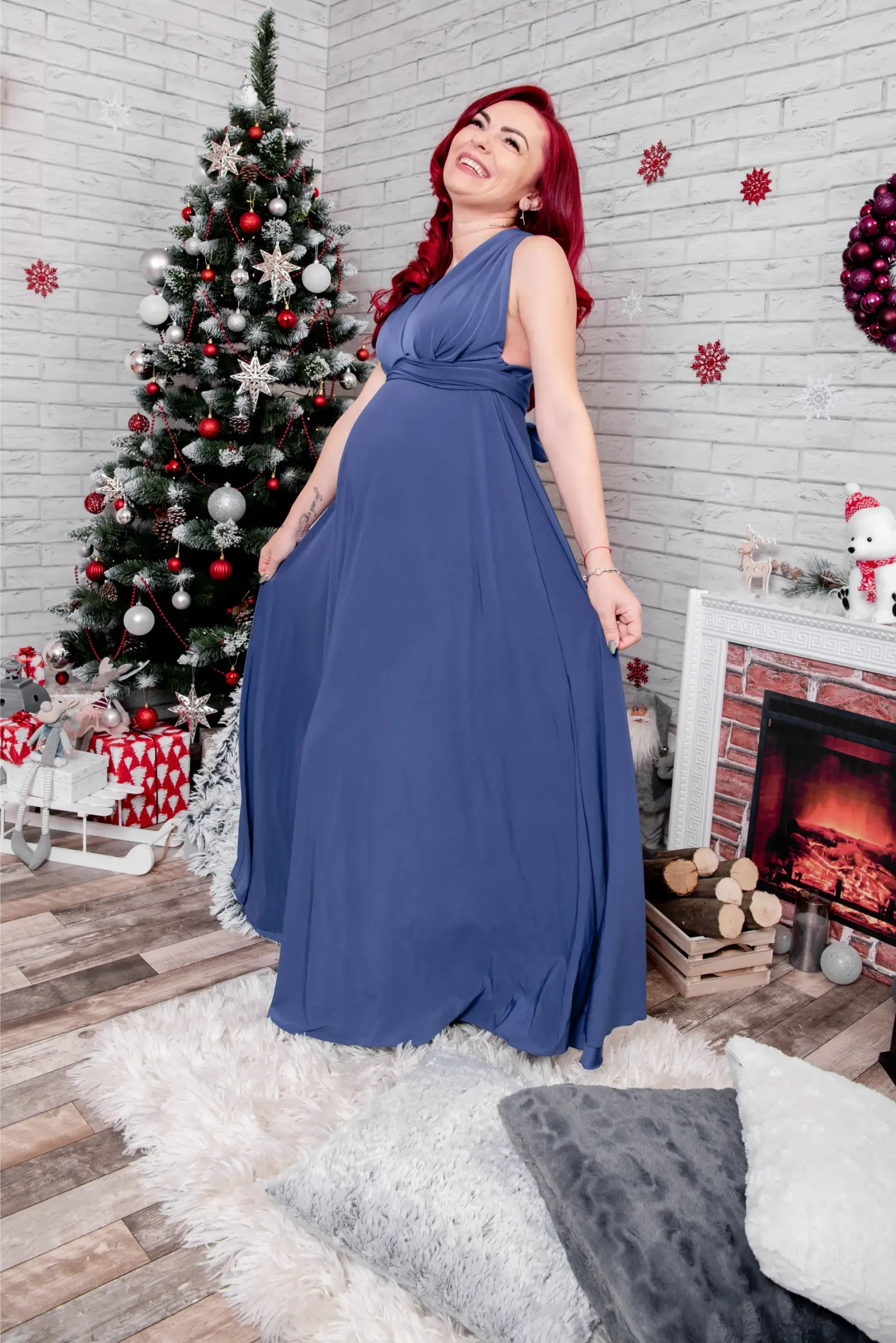 Mujer con vestido azul posando junto a un árbol de Navidad decorado y una chimenea en una habitación festiva.