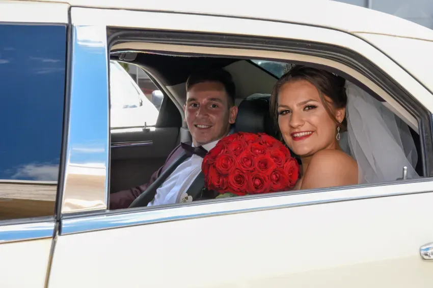 Pareja sonriendo en un coche blanco, con un ramo de rosas rojas en primer plano.