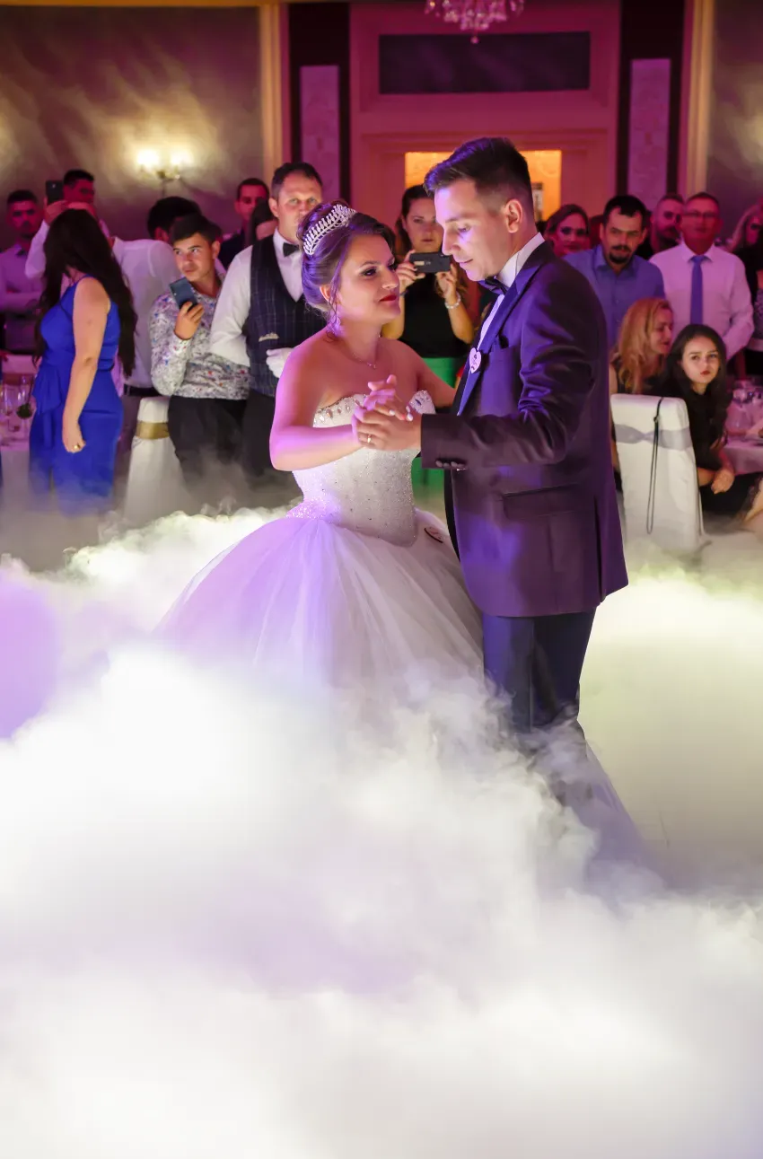 Los novios bailan entre la niebla baja durante una recepción nupcial, mientras los invitados observan de cerca.