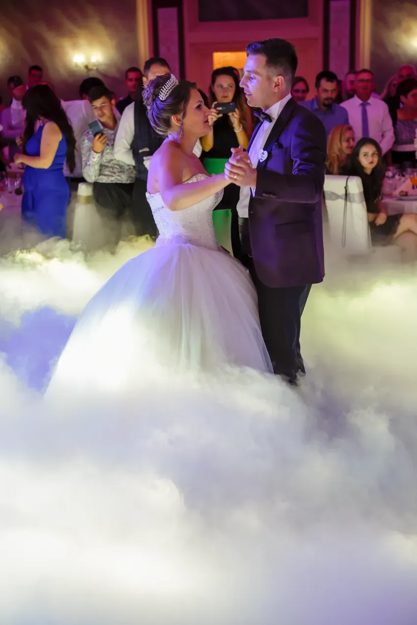 Novios bailando en un salón de banquetes lleno de niebla bajo una cálida iluminación rosa.