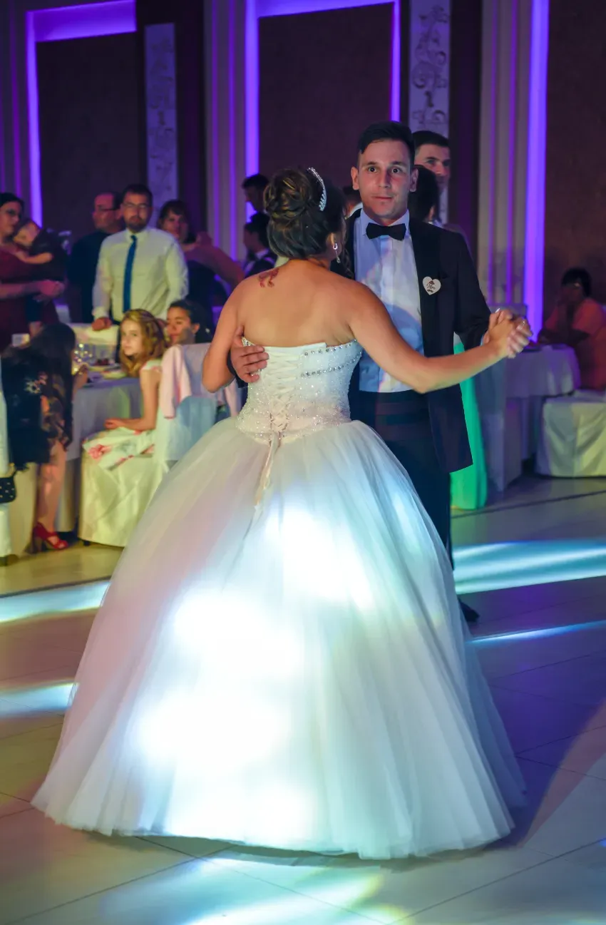 Los novios bailan lentamente en una recepción formal, su vestido blanco brilla bajo luces de colores.