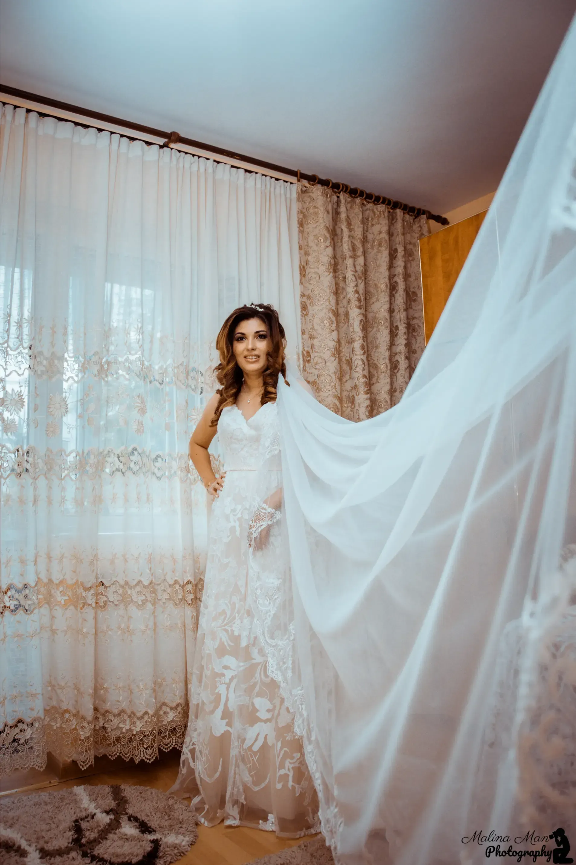 Novia con vestido de encaje posando en el interior junto a cortinas blancas transparentes y un velo vaporoso.