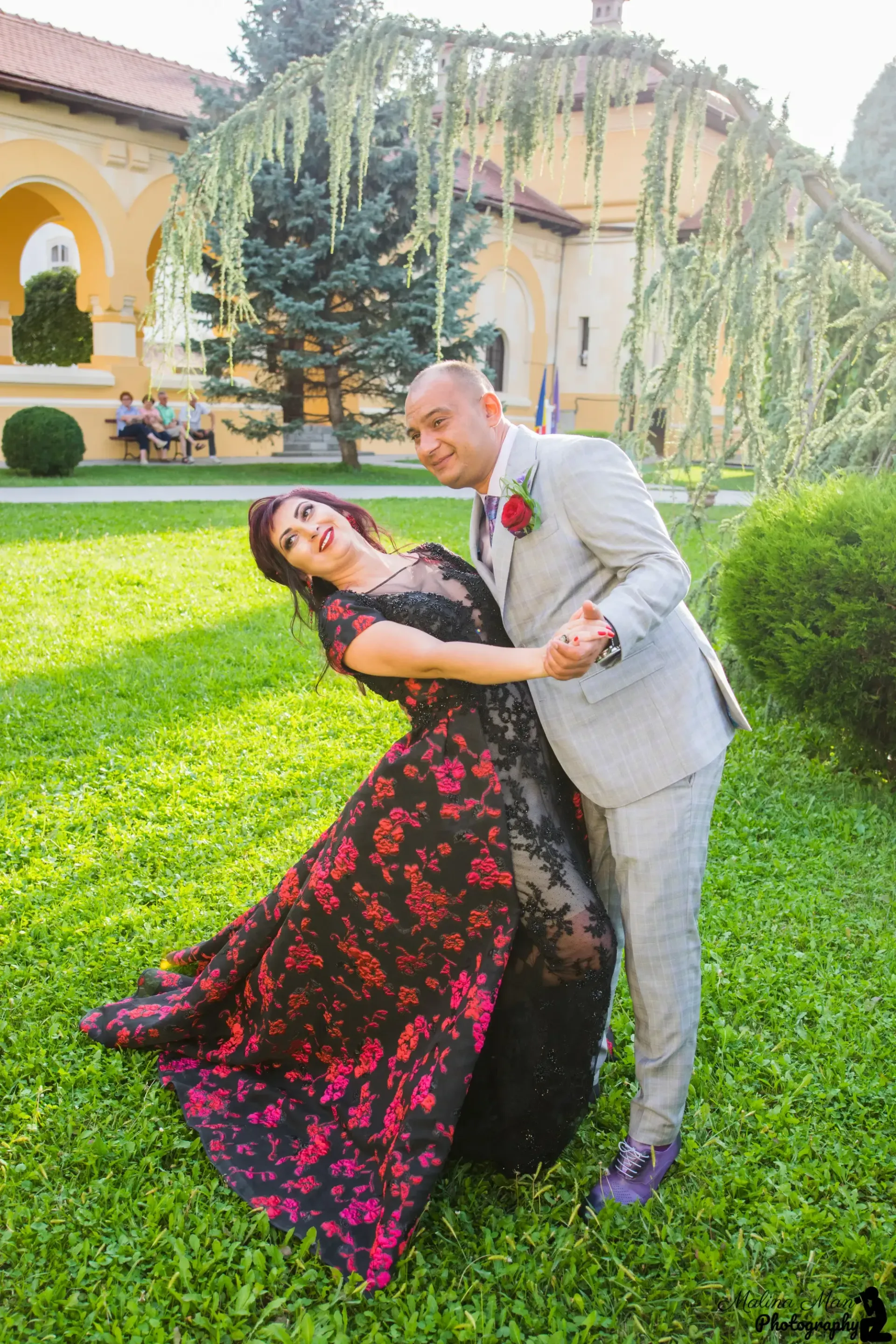Una pareja baila en el césped; la mujer lleva un vestido negro estampado de flores y el hombre un traje claro, sonriendo en un jardín.