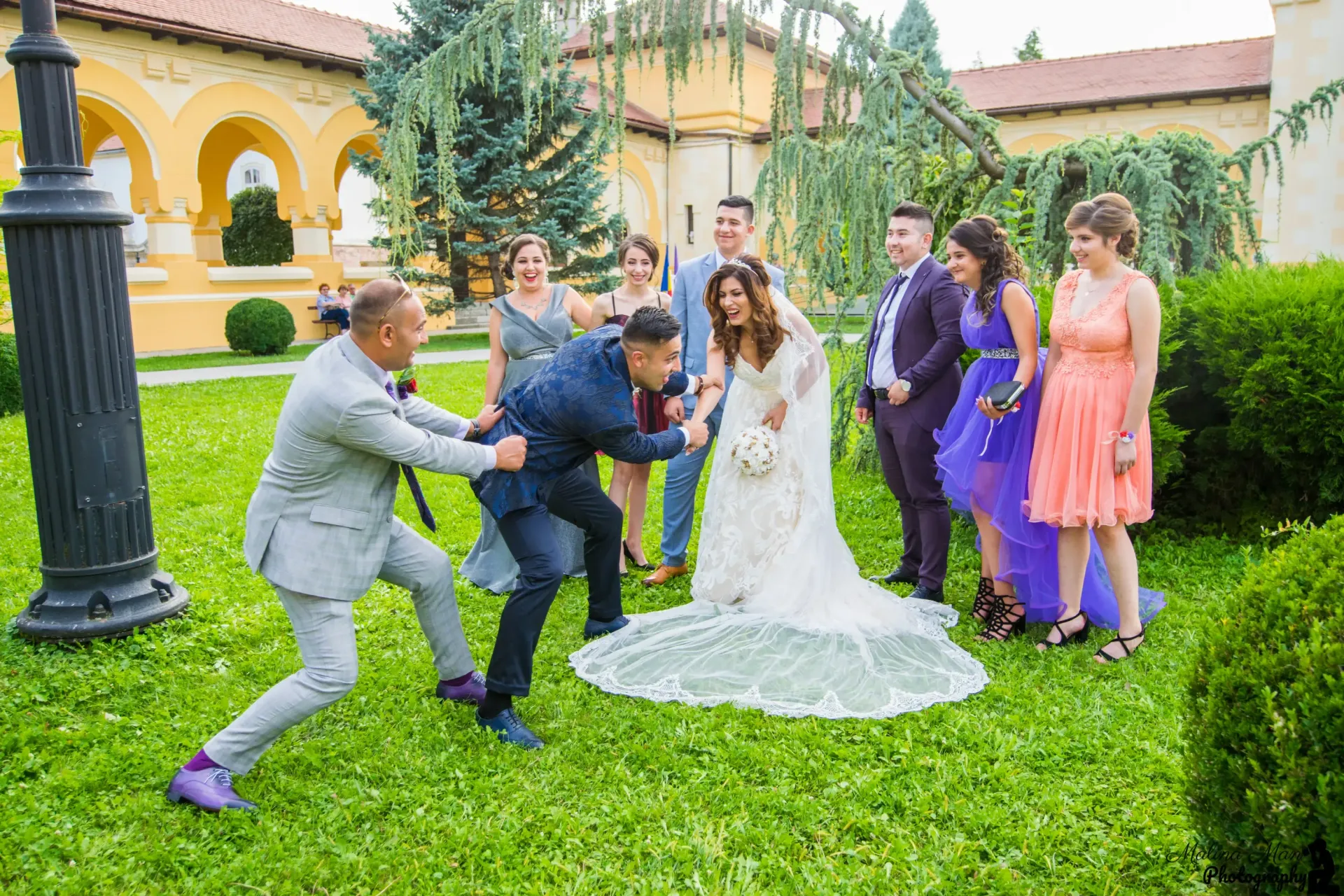Los invitados a la boda posan de forma divertida en el césped, con la novia en el centro, vestida con un vestido blanco, y los invitados con atuendos coloridos.