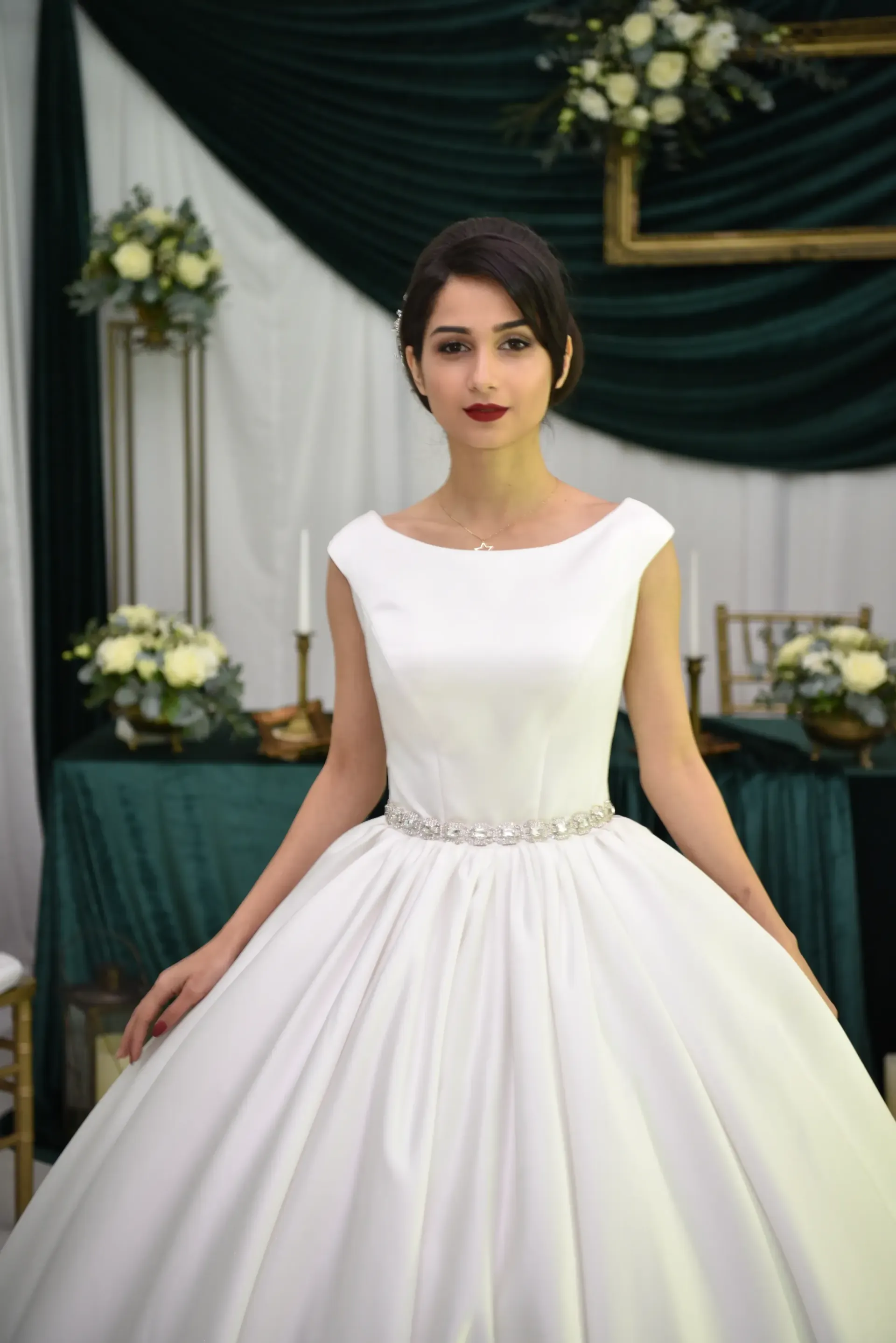 Mujer con un vestido blanco de pie frente a cortinas oscuras y decoraciones florales en un evento formal.