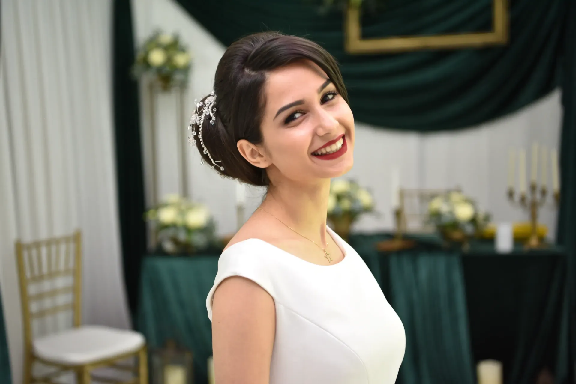 Mujer sonriente con vestido blanco en una mesa de evento decorada con cortinas verde oscuro.