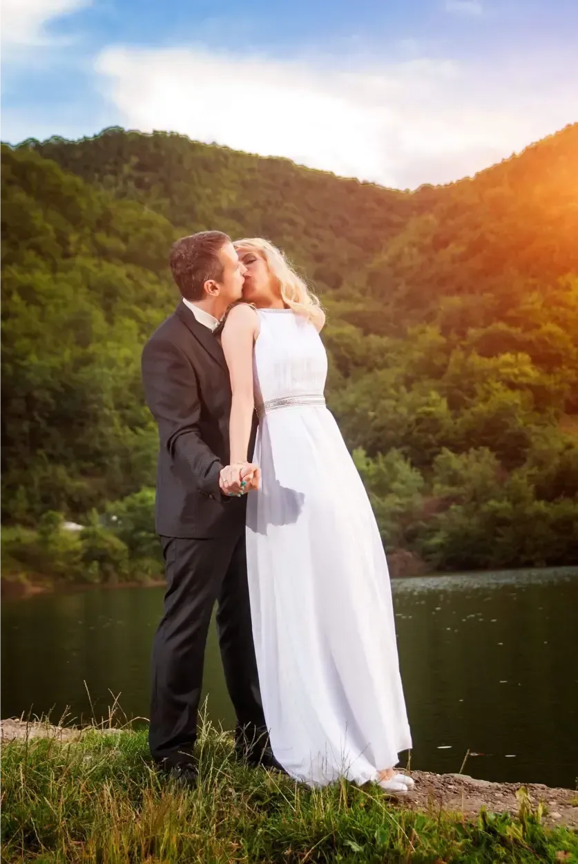Una pareja vestida de gala se besa junto a un lago al atardecer, con verdes colinas de fondo.