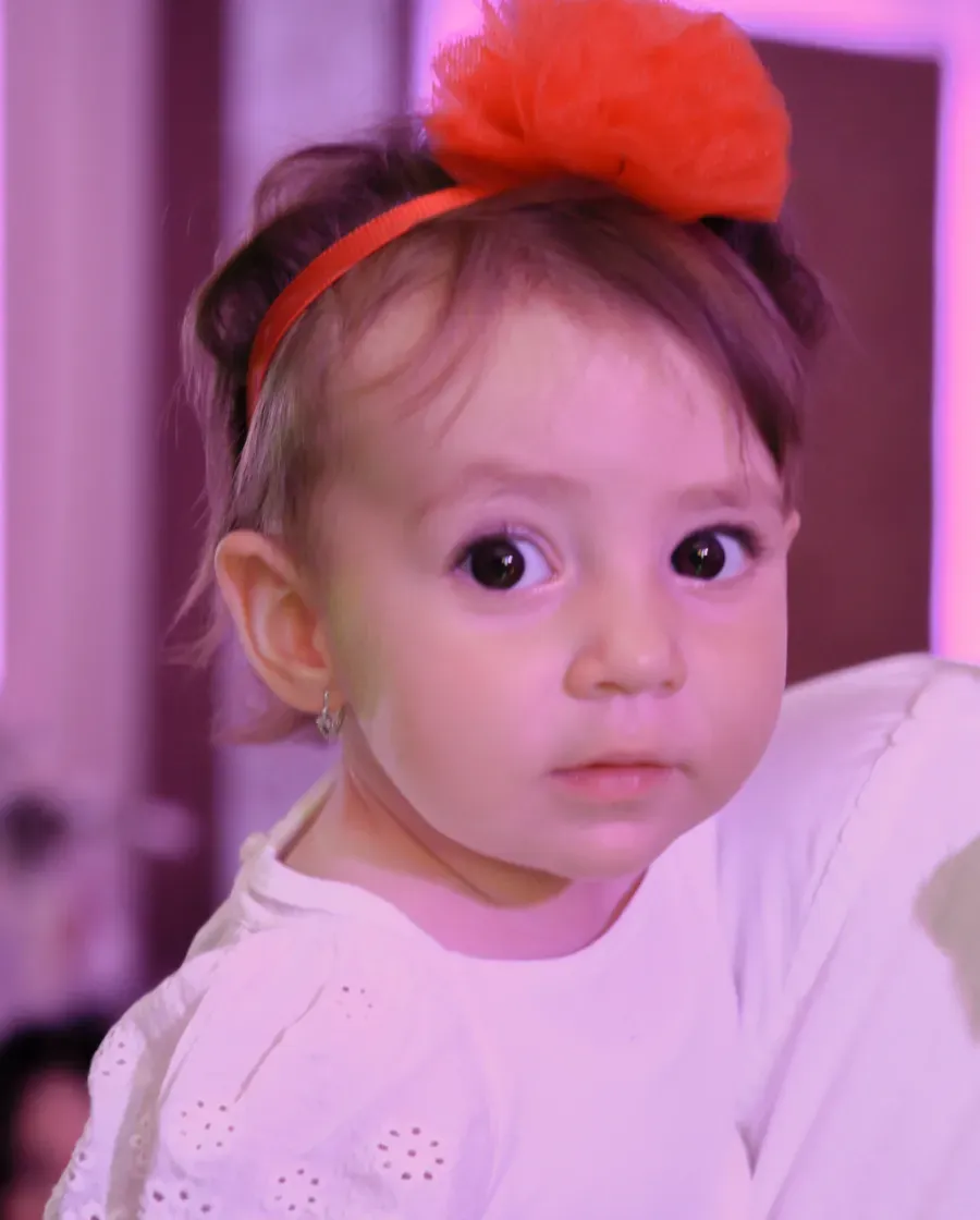 Una niña pequeña con un lazo rojo mira a la cámara sobre un fondo rosa suavemente iluminado.