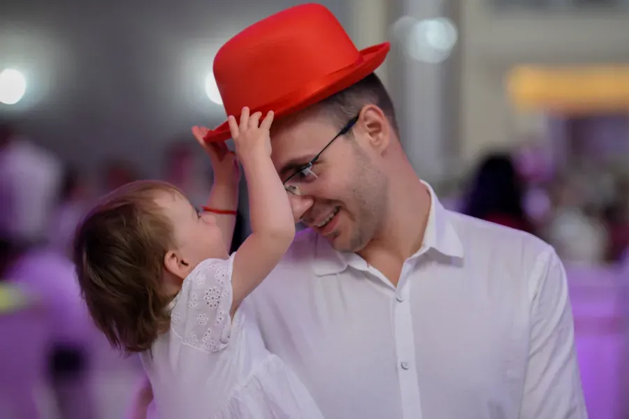 Un niño le ajusta un sombrero rojo brillante a un hombre sonriente con camisa blanca, en un interior con luces borrosas.