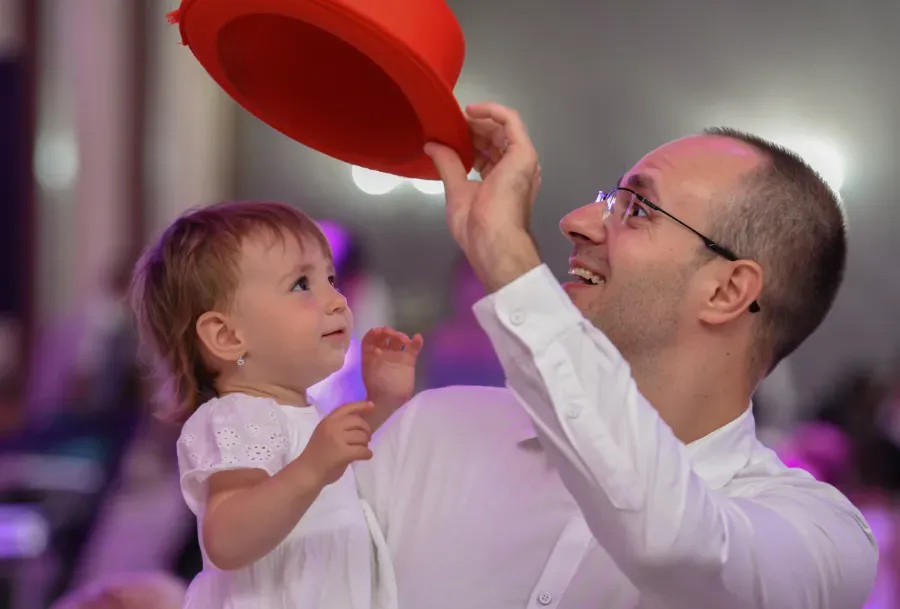 Un hombre y un niño con camisas blancas sonríen y extienden la mano hacia un globo rojo en un luminoso entorno interior.