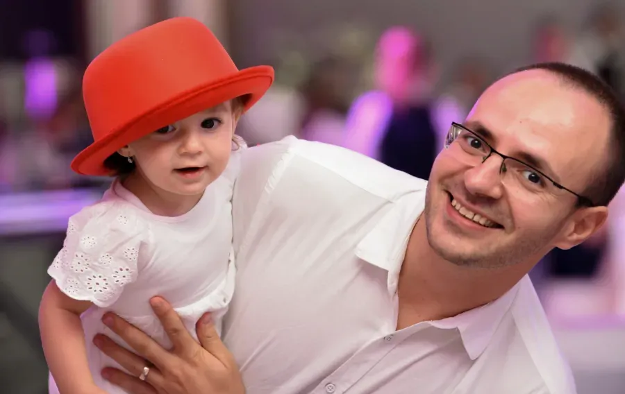 Un adulto sostiene a un niño con un sombrero rojo; ambos sonríen en un evento festivo con luces moradas borrosas al fondo.