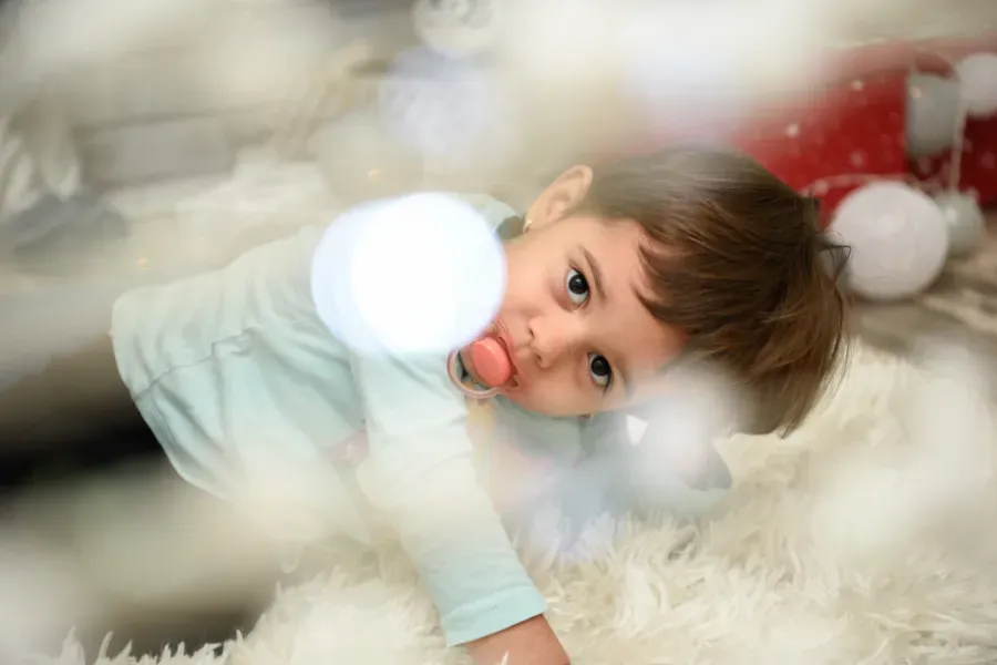 Un niño yace sobre una alfombra blanca y mullida, mirando a la cámara con luces suaves y borrosas de fondo.