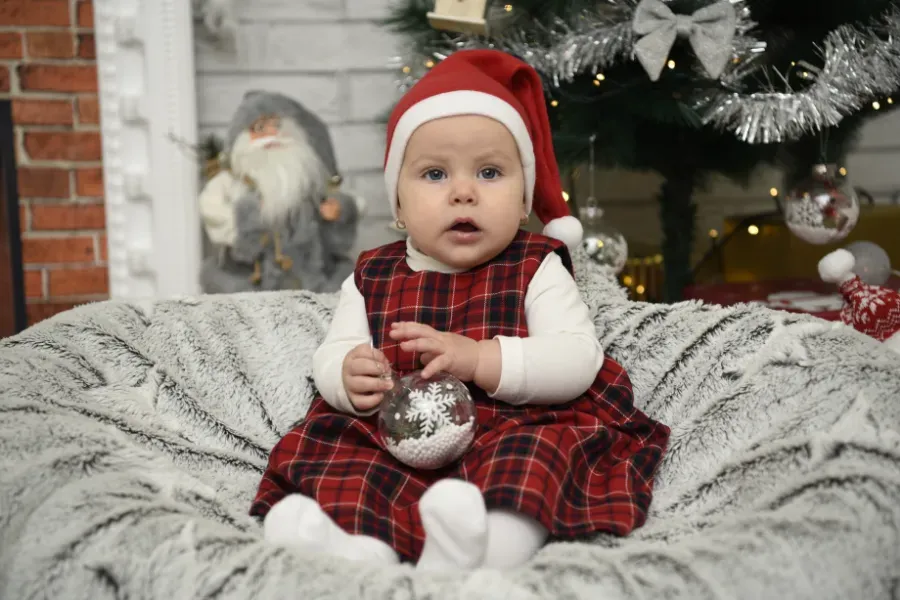 Bebé con un vestido de cuadros rojos y gorro de Papá Noel sentado junto a un árbol de Navidad decorado, sosteniendo un adorno plateado.