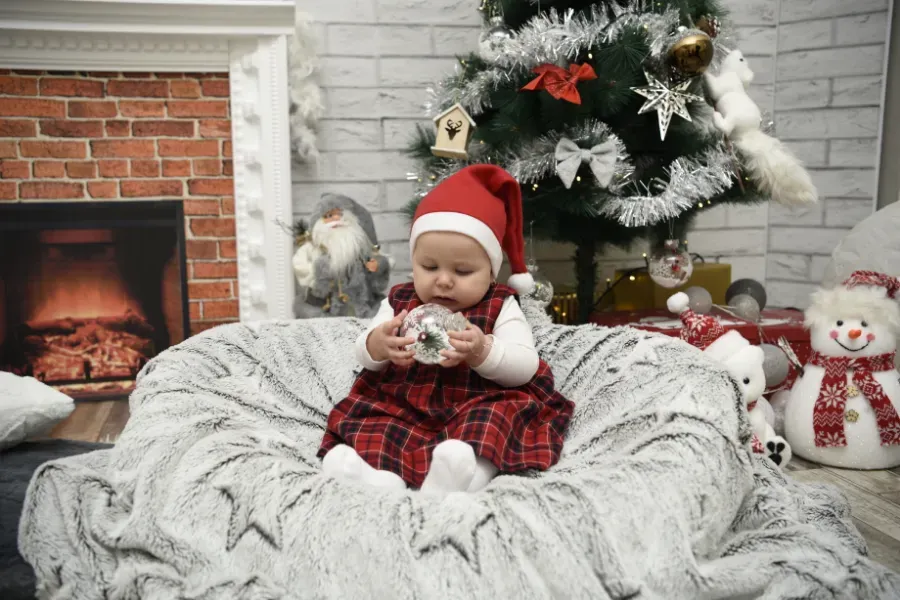Un bebé vestido de Papá Noel está sentado en un puf gris junto a un árbol de Navidad y una chimenea.