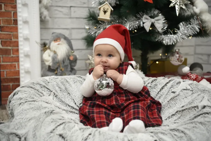 Bebé con vestido de cuadros rojos y gorro de Papá Noel sosteniendo un adorno plateado sobre una manta con motivos invernales.
